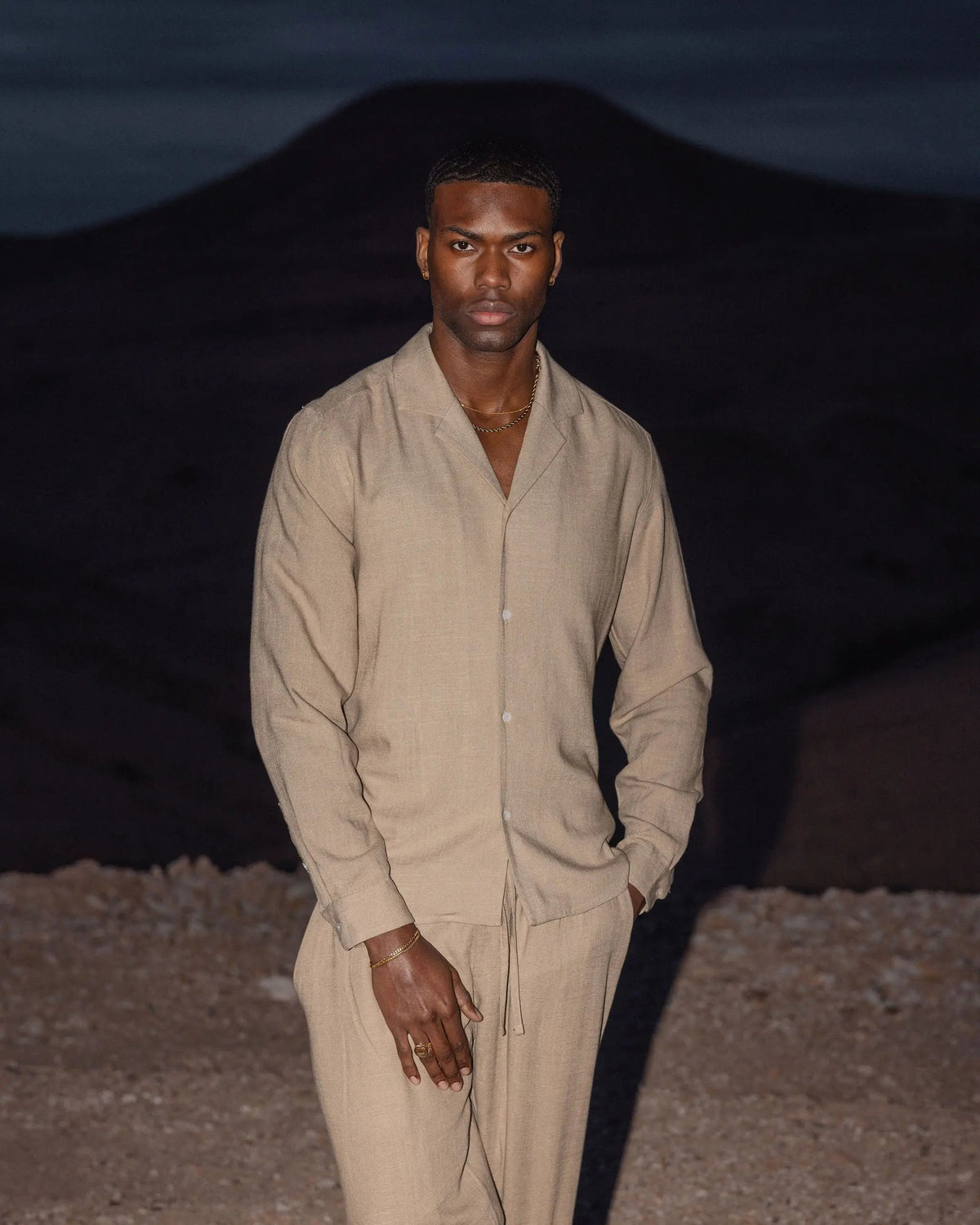 Homme debout dans un paysage désertique au crépuscule, portant une chemise en lin à col ouvert et un pantalon assorti dans un beige sable