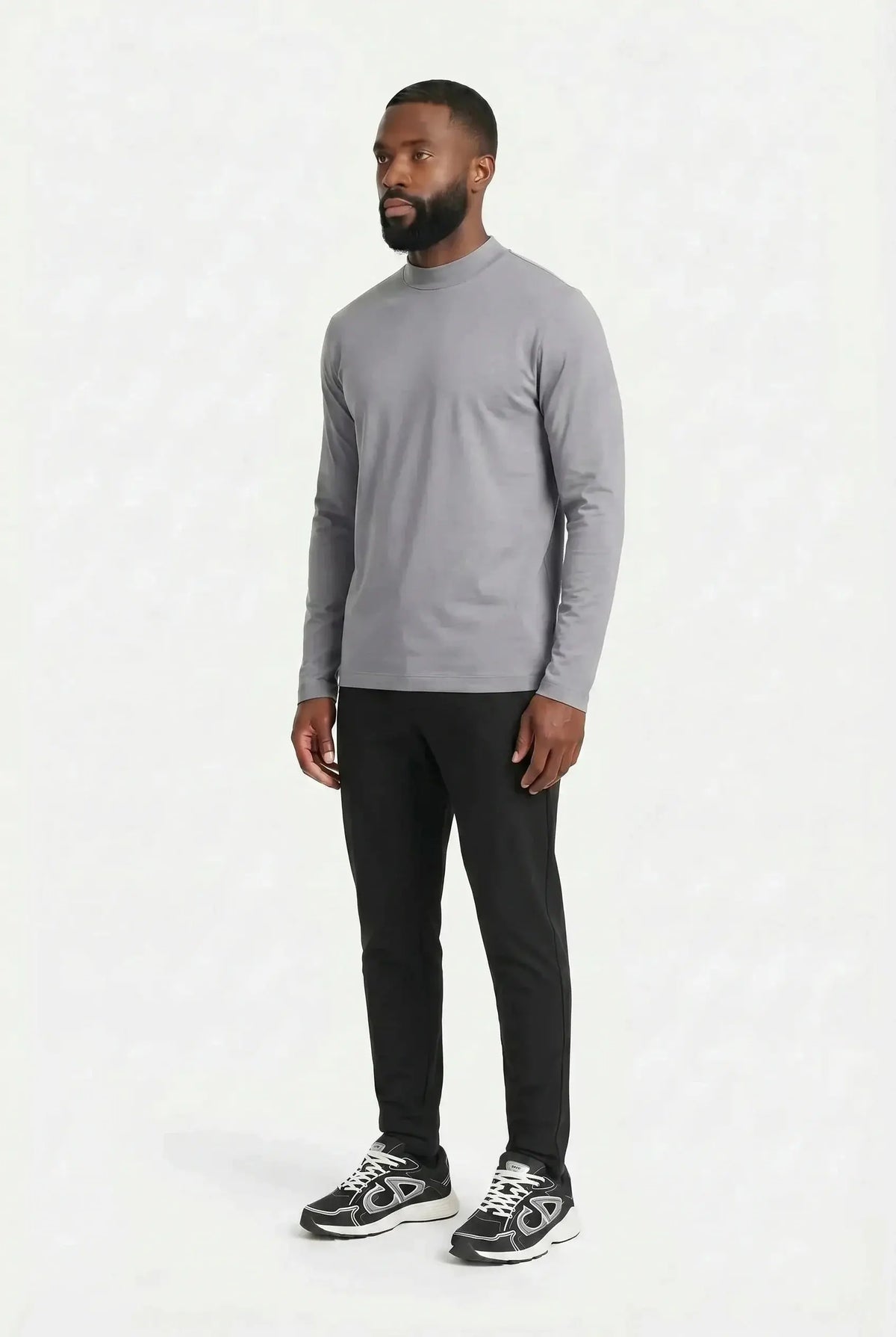 Homme debout de trois quarts, silhouette entière, en t-shirt à manches longues gris perle et pantalon noir, sur fond clair uni